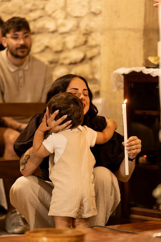 L'enfant baptisé, de dos, dans les bras de sa maman qui tient un cierge allumé